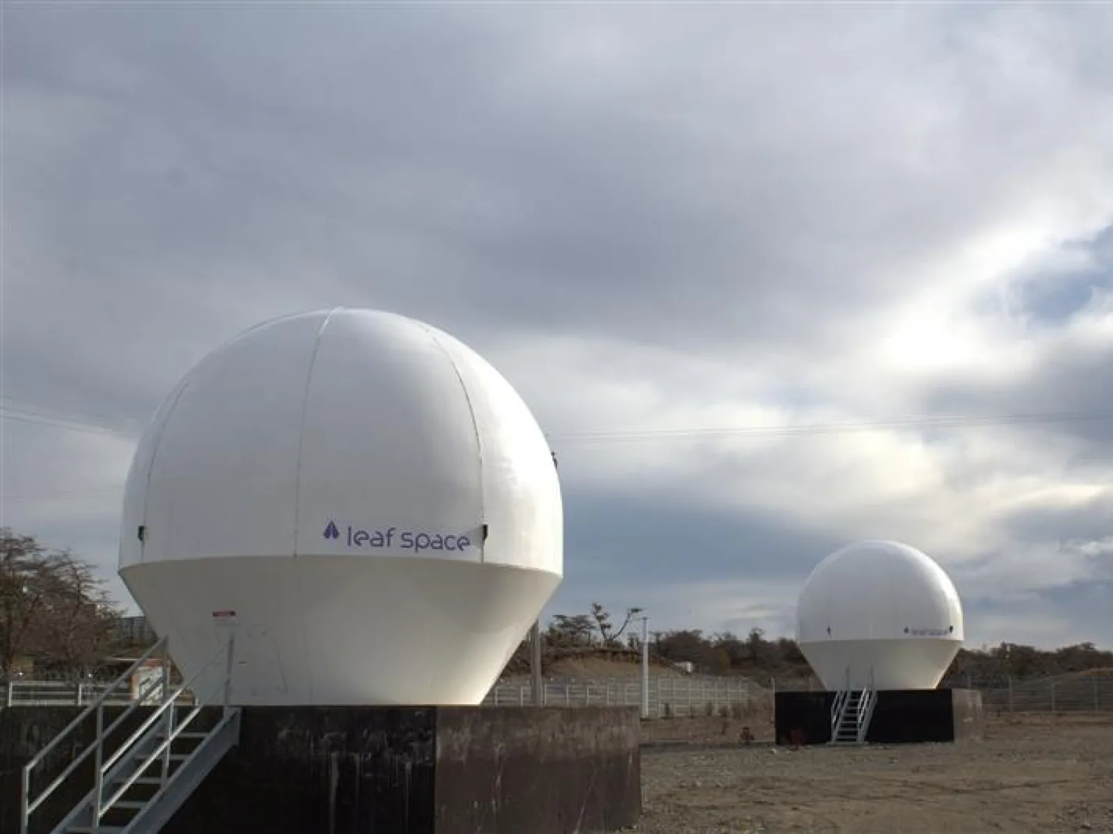 Two white Leaf Space ground station antenna radomes on raised platforms under a cloudy sky