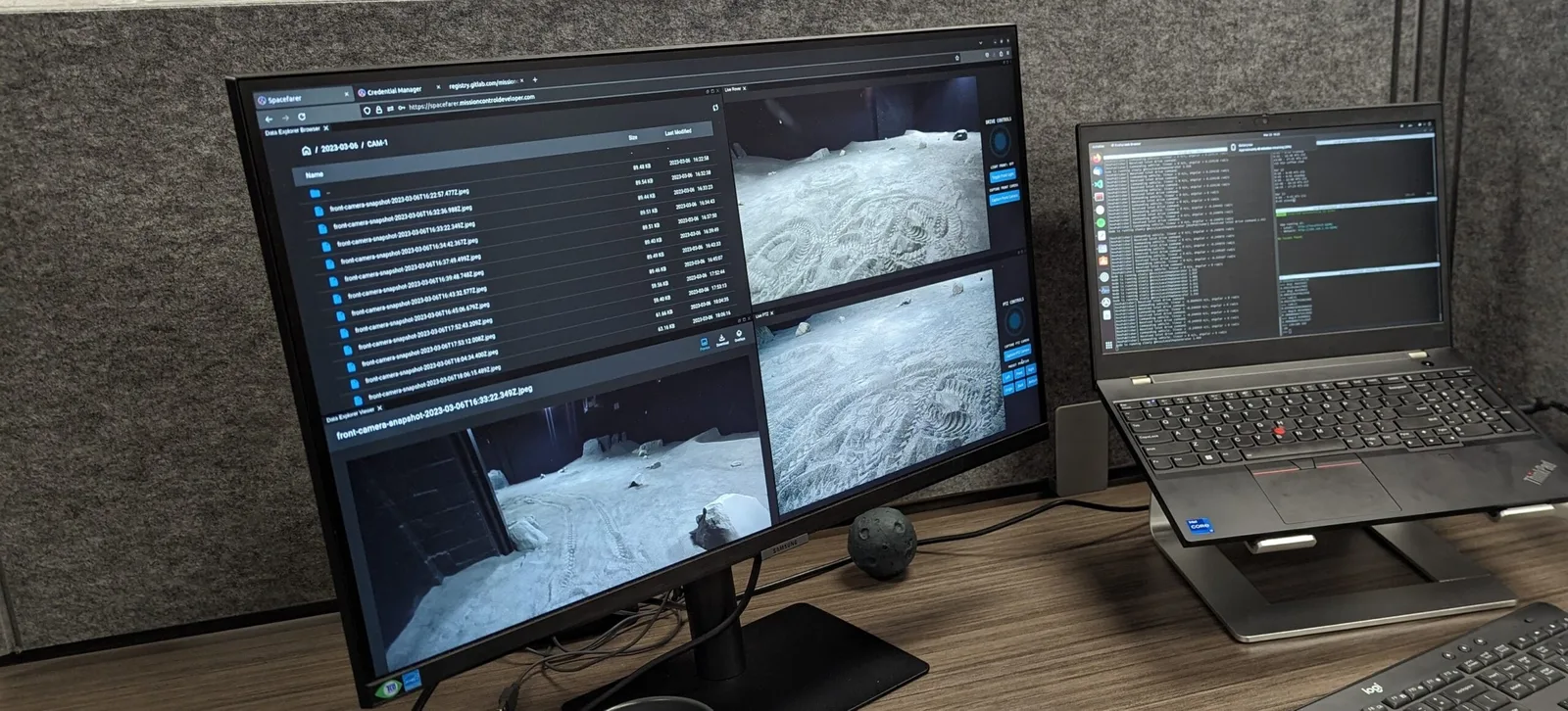 Monitor running Mission Control's Spacefarer software with rover camera feeds of a simulated lunar surface, beside a laptop