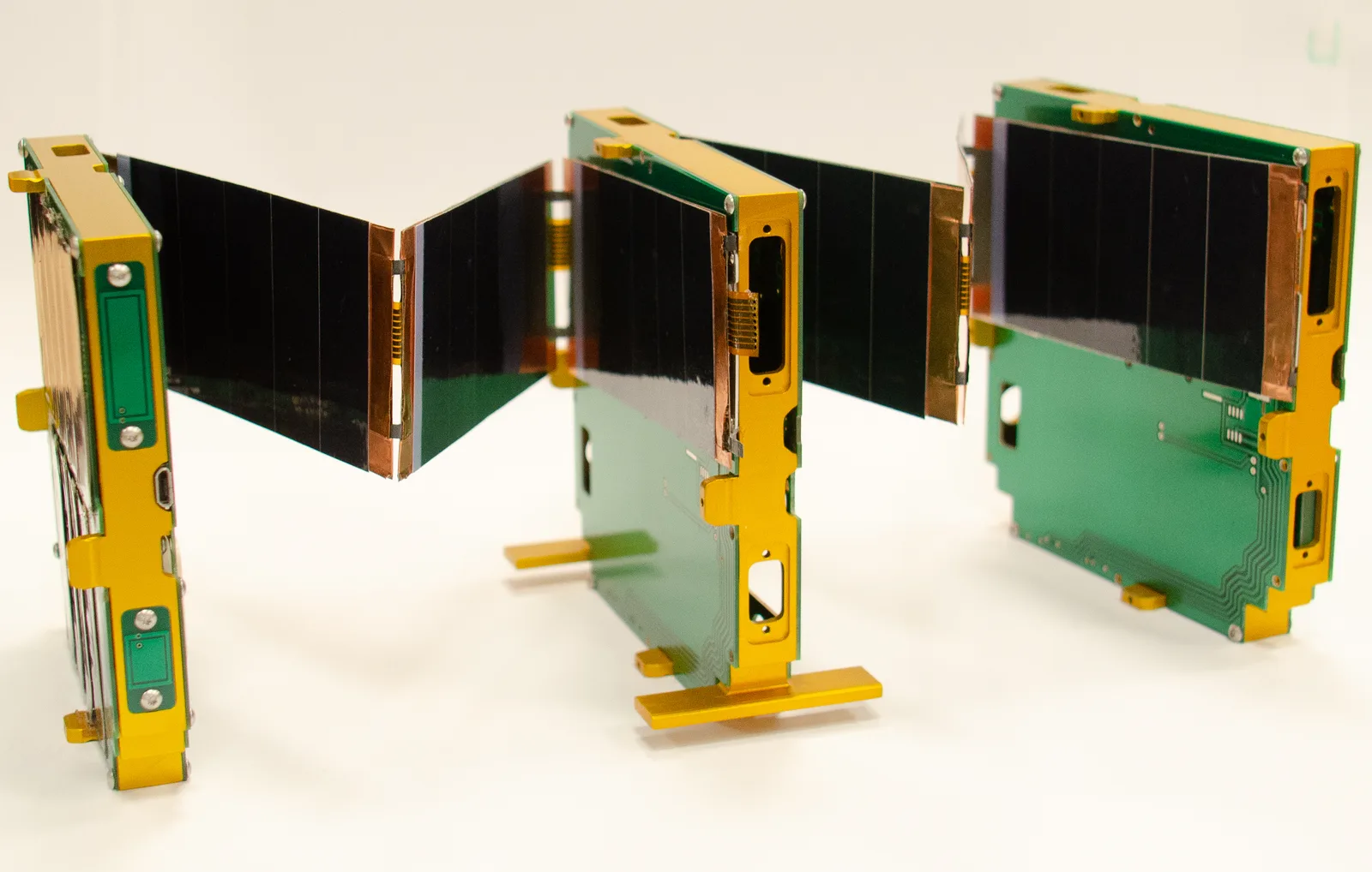 Three linked ThinSat picosatellites with gold frames and solar panels standing on a white surface