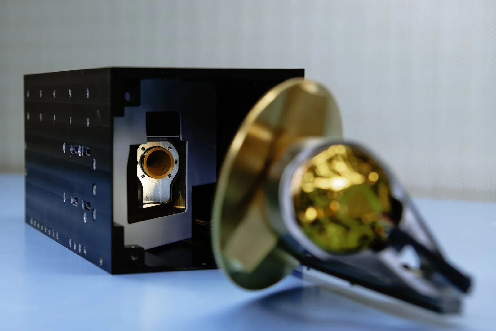 A GEMS microwave weather satellite on a bench, showing its black chassis and gold radiometer antenna